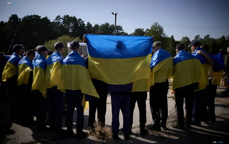 Group of people draped in Ukrainian flags stands back-to-camera holding a large Ukrainian flag — symbolizing hope for prisoner exchange