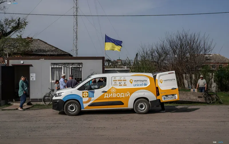 LKW der Ukrposhta mit UNICEF-Logo und der Aufschrift «Dyvodi» steht vor einem ländlichen Postamt, wo Menschen Pakete erhalten. Im Hintergrund die ukrainische Flagge und eine ländliche Gegend.