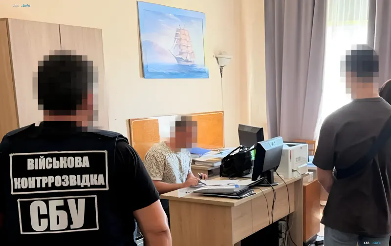 Співробітник СБУ в жилеті з написом 'Військова контррозвідка СБУ' проводить допит в кабінеті, де сидить затриманий доцент Міноборони України, звинувачений у шпигунстві на користь ГРУ та злитті креслень дронів.