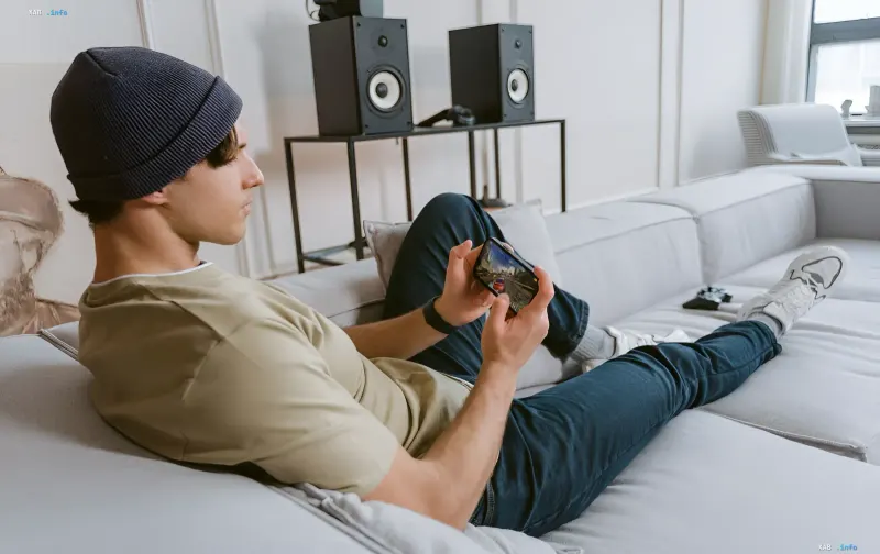 A young man in a beanie plays a mobile game on a smartphone while lying on a sofa in a modern living room