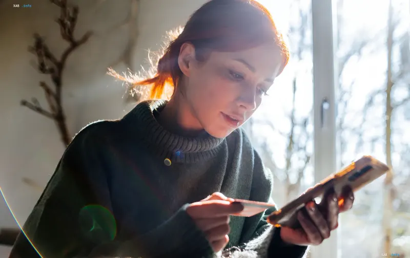 Frau in gemütlichem Pullover nutzt Smartphone und hält Bankkarte, um Ratenzahlung zu planen – Illustration zum Artikel über finanzielle Kompetenz der Ukrainer