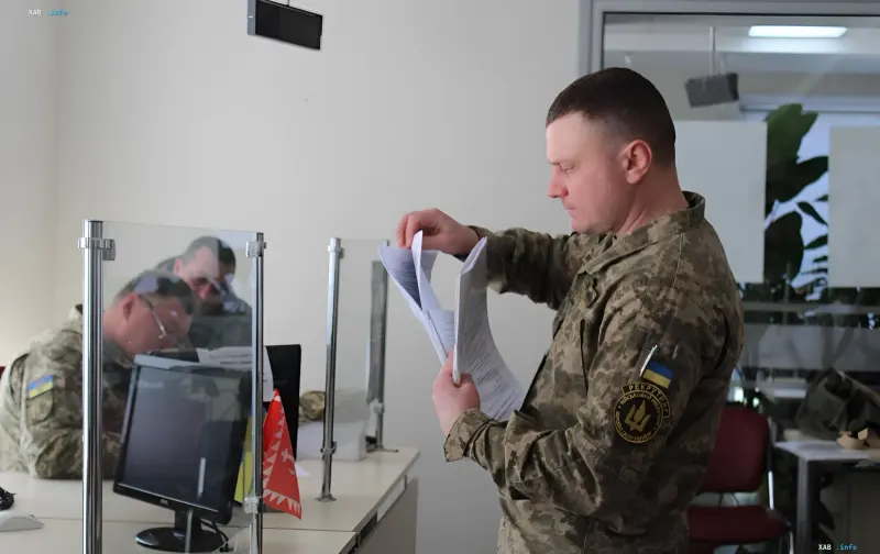 Украинский военный в камуфляже изучает документы в офисе ТЦК, символизируя продление мобилизации и военного положения до 2026 года