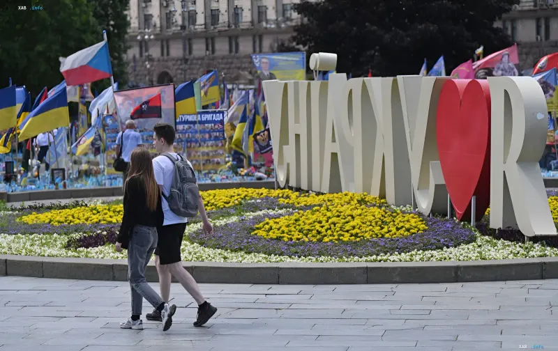 Молодая пара гуляет по солнечному парку в Киеве, рядом с цветочной клумбой и памятником «Я люблю Киев», на фоне украинских флагов — символ возвращения жары и летнего настроения в Украине