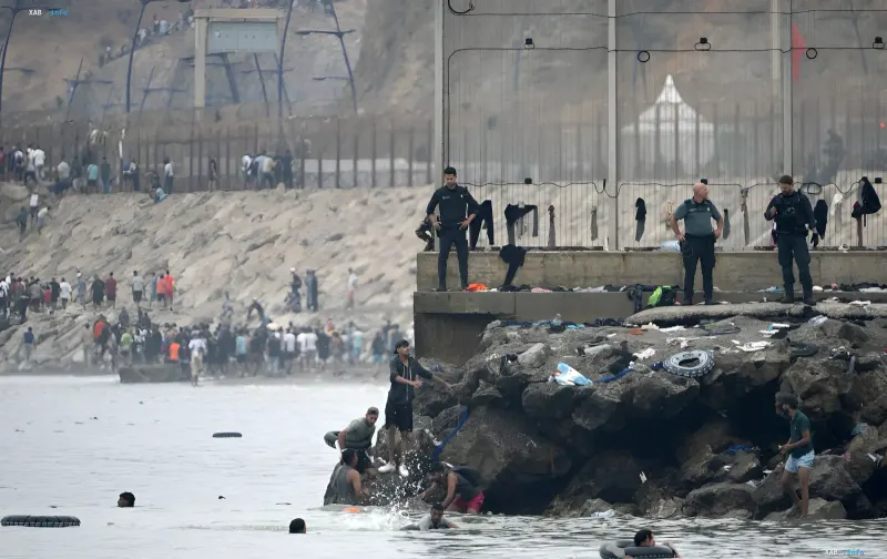 Migranten schwimmen über das Meer in der Nähe europäischer Küsten, während Grenzbeamte von einer Betonplattform aus beobachten – eine Illustration der Migrationskrise, auf die Frankreich mit verstärkten Maßnahmen reagiert