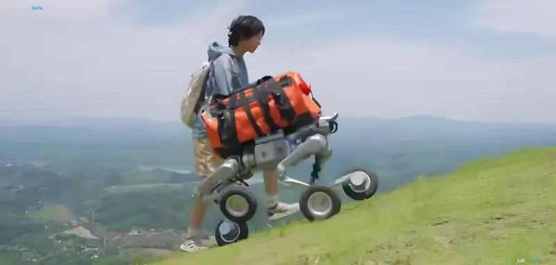 Person nutzt den Unitree AS2-W-Roboter, um einen schweren orangefarbenen Rucksack einen steilen grasbewachsenen Hang hinauf zu ziehen und demonstriert die tatsächliche Tragfähigkeit der Plattform.