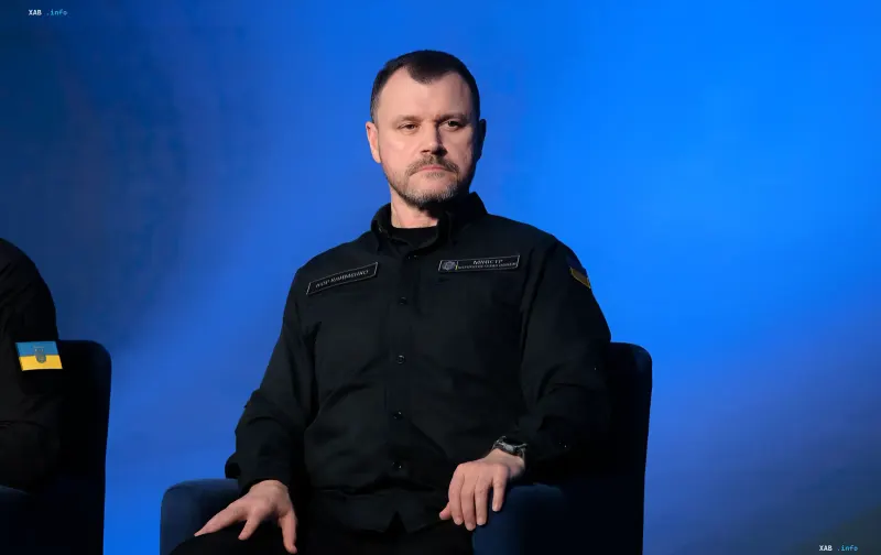 Ihor Klymenko in schwarzer Uniform mit ukrainischer Flagge vor einem blauen Bildschirmhintergrund, der seine neue Rolle in der NSDC symbolisiert