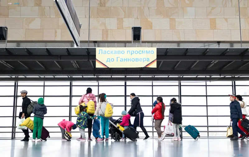 Группа украинских беженцев с чемоданами и сумками стоит у выхода на вокзале в Германии под вывеской 'Ласкаво просимо до Ганновера'