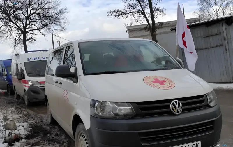 Белый фургон Volkswagen с эмблемой Украинского Красного Креста и красным крестом на капоте, припаркованный рядом с другим гуманитарным транспортом на фоне зимнего пейзажа.