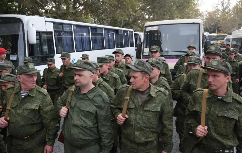 Группа российских военных в камуфляжной форме и фуражках с эмблемами, идущих рядом с белыми автобусами, иллюстрируя тему скрытых ограничений на выезд мужчин призывного возраста в России.