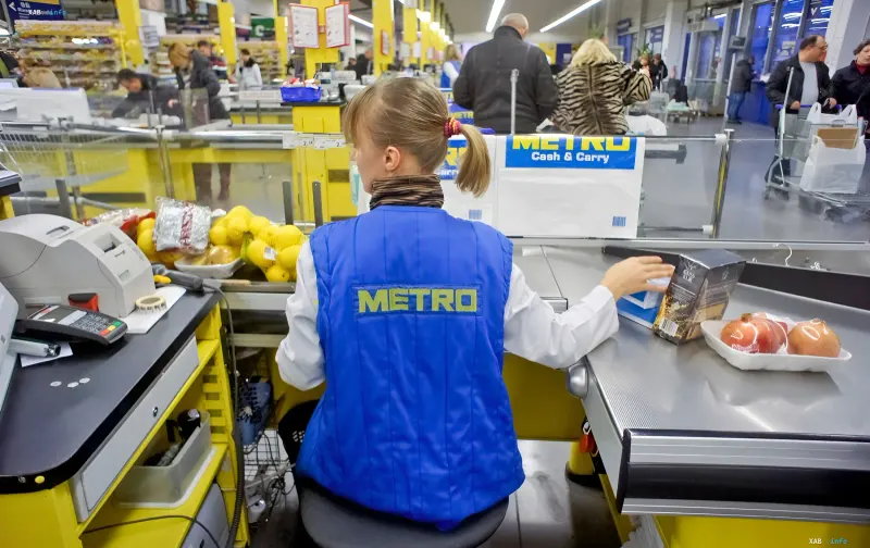 Кассир в синем жилете Metro за кассой Cash & Carry сканирует товары на фоне очереди покупателей в супермаркете