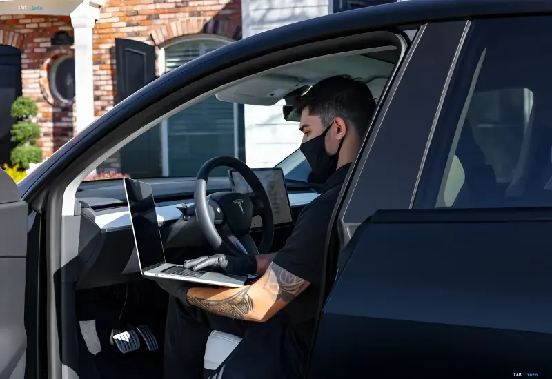 Fahrer mit Maske und Handschuhen arbeitet im Tesla-Interieur an einem Laptop, während die Innenraumkamera seine Handlungen aufzeichnet