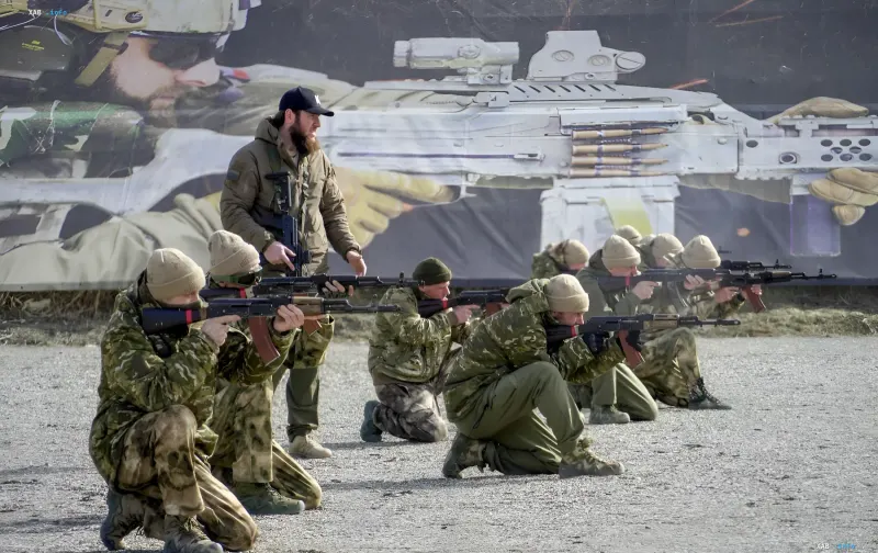 Russian MoD recruitment event: cadets in camouflage aim rifles under an instructor's supervision against a banner of military hardware