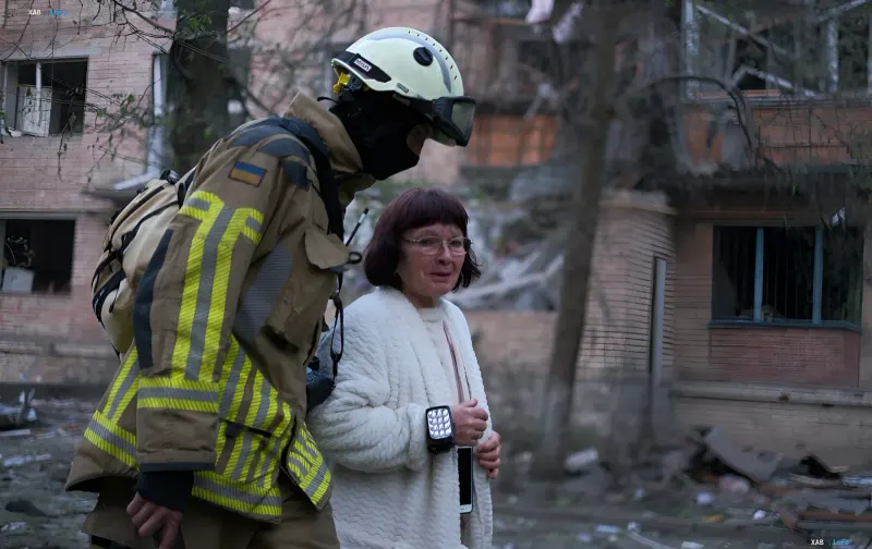 Рятувальник у захисному спорядженні з прапором України допомагає похилій жінці на тлі зруйнованого будинку після ракетного удару — ілюстрація дефіциту ППО «Патріот»
