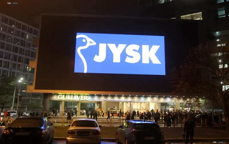 Нічний фасад магазину JYSK в Україні з великим логотипом на екрані та натовпом покупців біля входу, мережа закриває точки при будь-якій повітряній тривозі