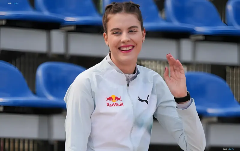Марта Магучих у білій куртці з логотипами Red Bull і Puma посміхається та махає рукою на тлі трибун стадіону перед виходом на червону доріжку Абсолютного ЧМ у Будапешті