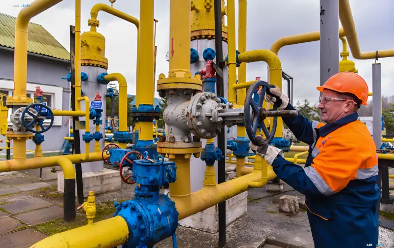 Работник в каске и защитных очках поворачивает вентиль на газовом объекте с желтыми трубами
