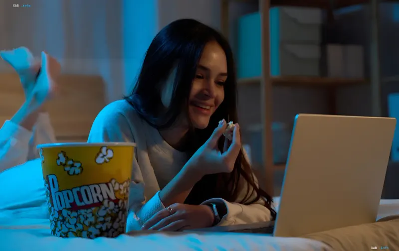 A young woman lying on a bed watching a series on her laptop and eating popcorn from a bucket at night.