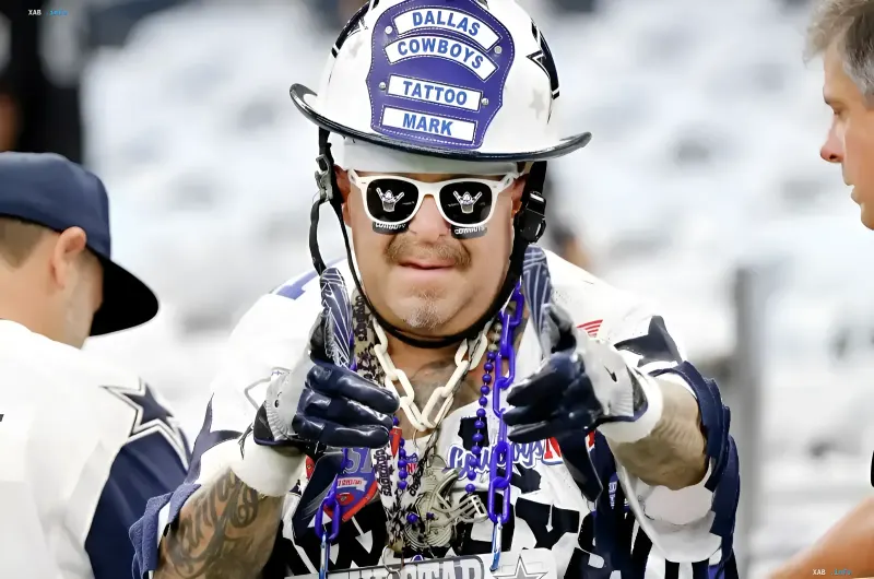 Мужчина в белом шлеме пожарного и солнечных очках с символикой Dallas Cowboys показывает пальцами жест на камеру.