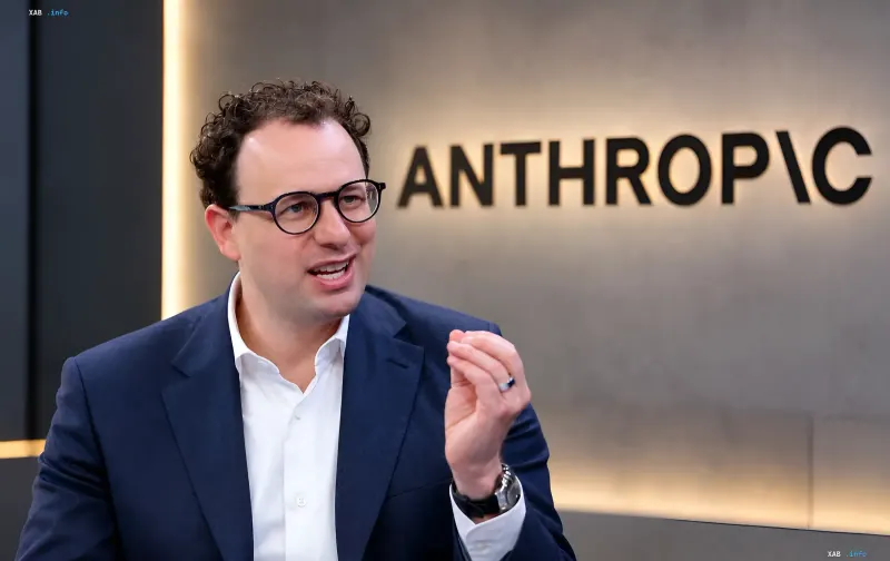 An Anthropic executive in a business suit gestures while speaking in front of the company logo