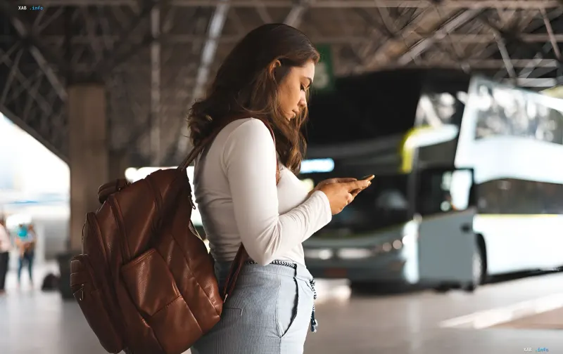 Woman with a backpack checking her phone at a bus station in front of a bus