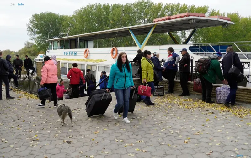 People with suitcases waiting by a river boat on the shore during a humanitarian evacuation