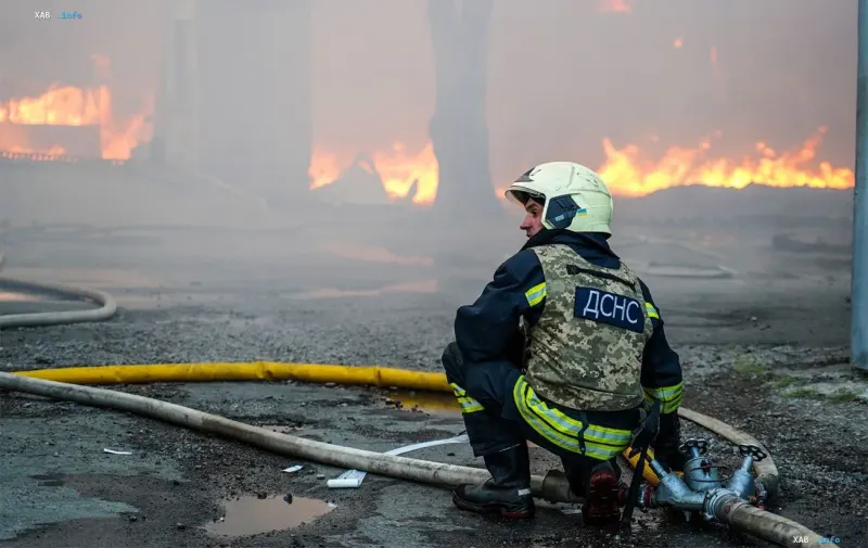 Рятувальник ДСНС у касці та захисному жилеті сидить на тлі масштабної пожежі після ракетного удару у Лозовій