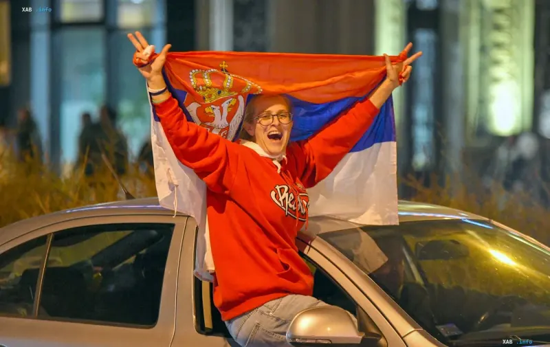 Женщина радостно машет сербским флагом, стоя в люке движущегося автомобиля в вечернее время