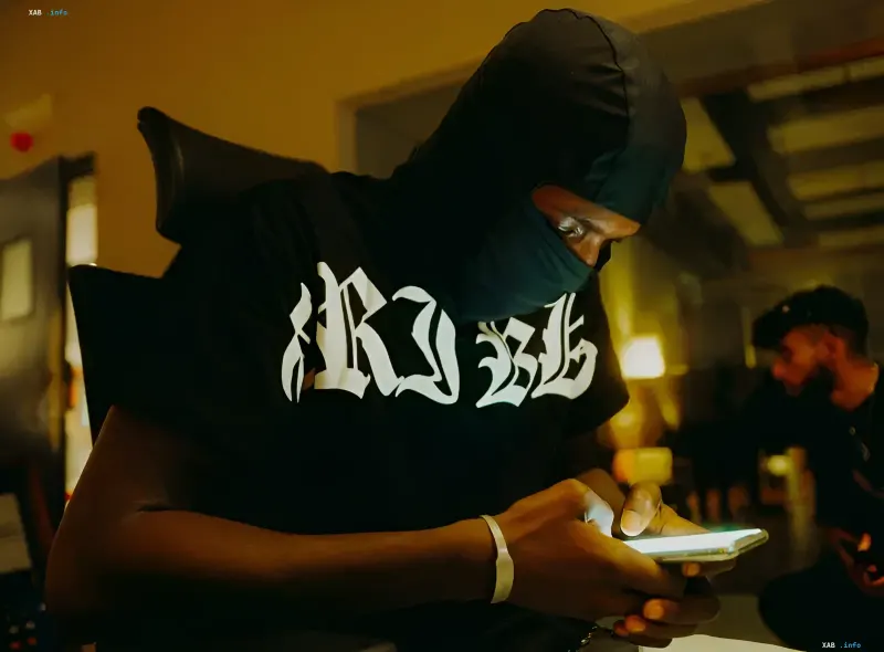 A person in a balaclava sits indoors checking their smartphone under yellow lighting