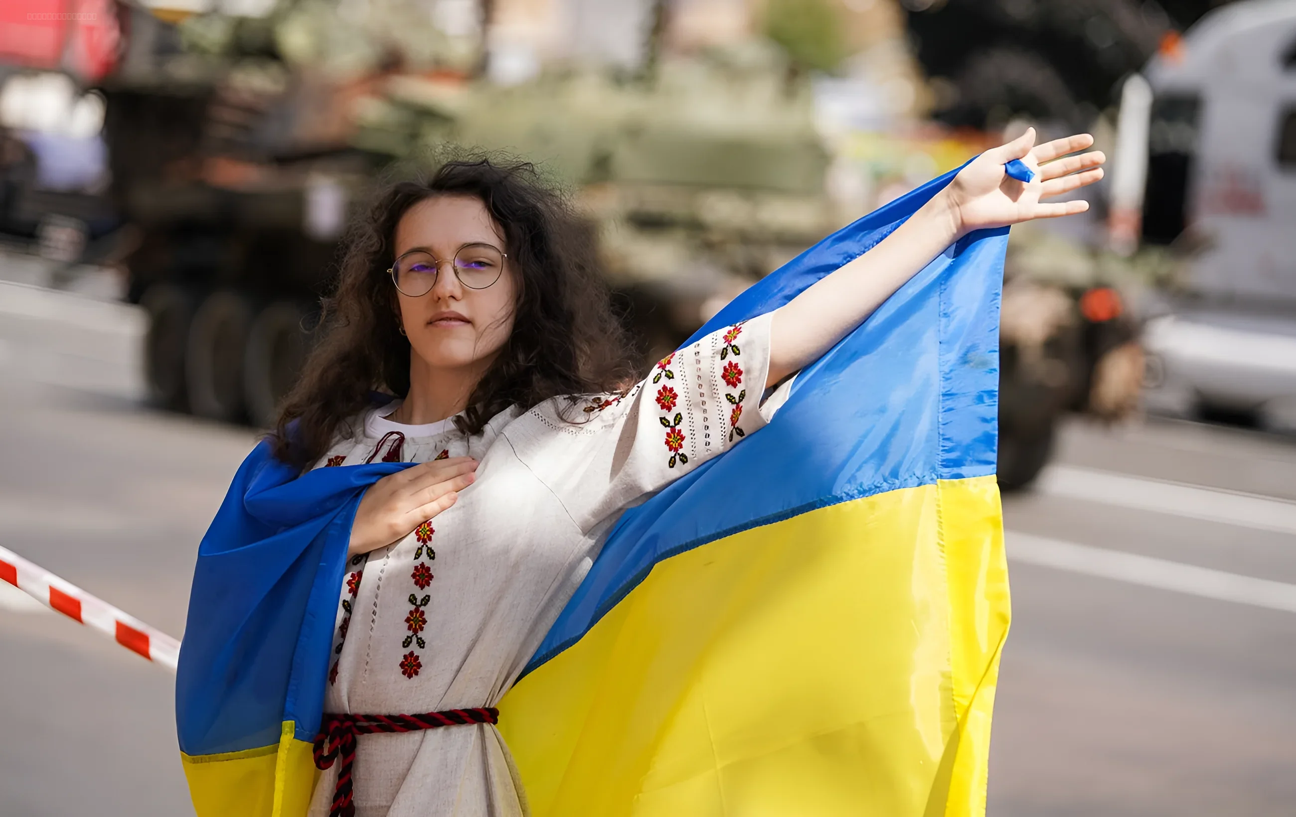 Девушка в традиционной вышиванке держит украинский флаг на фоне военной техники — символ культурного сопротивления и патриотизма в День украинской музыки