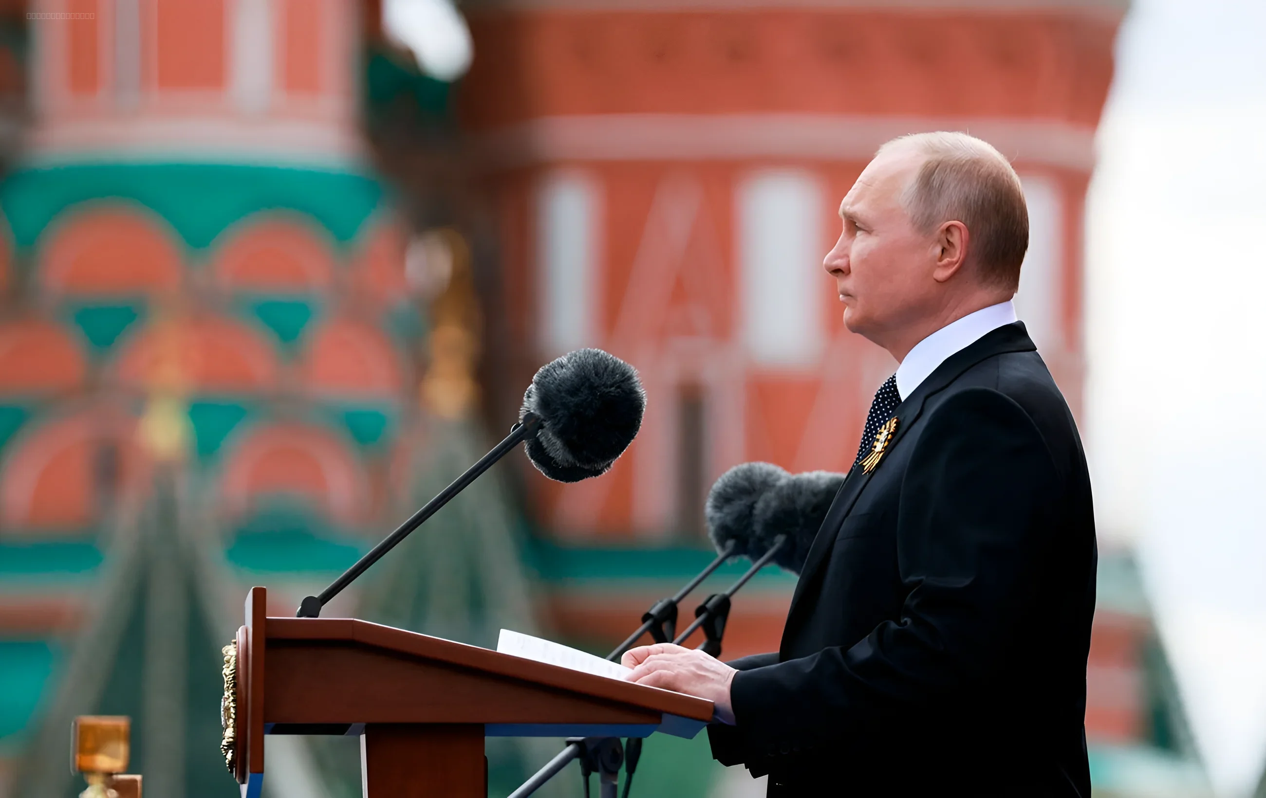 Vladimir Putin speaking at a podium during a military parade, with blurred red Kremlin walls in the background