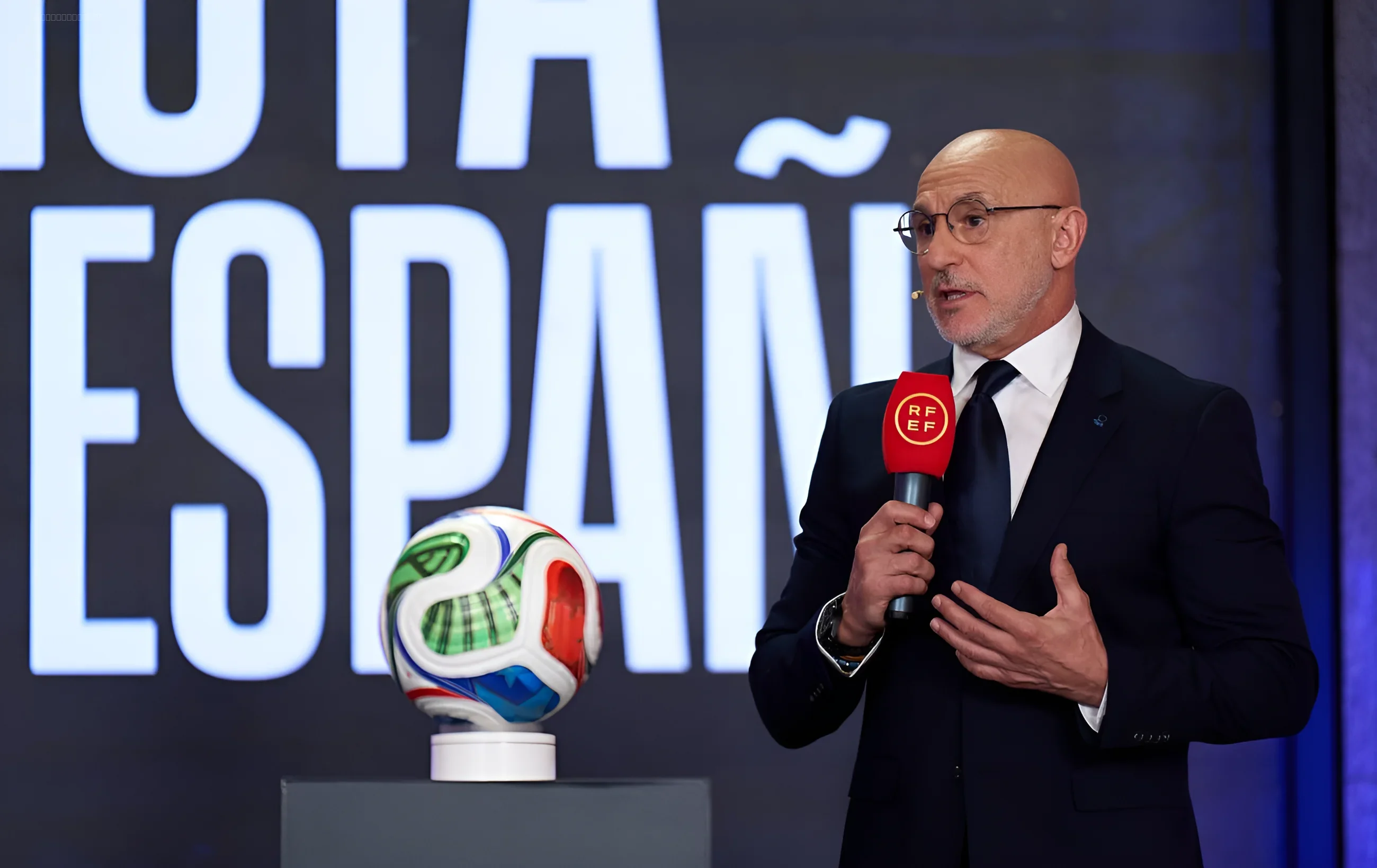 Луис де ла Фуэнте с микрофоном RFEF на фоне футбольного мяча и надписи 'ESPAÑA'