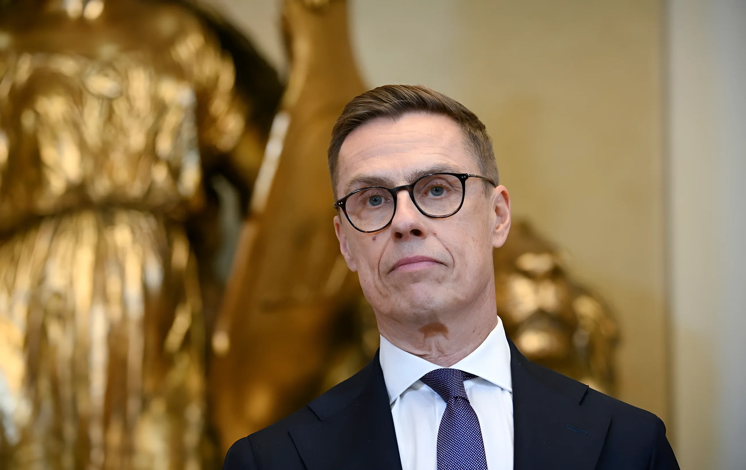 Президент Финляндии Александр Стубб в очках и костюме на фоне золотой статуи