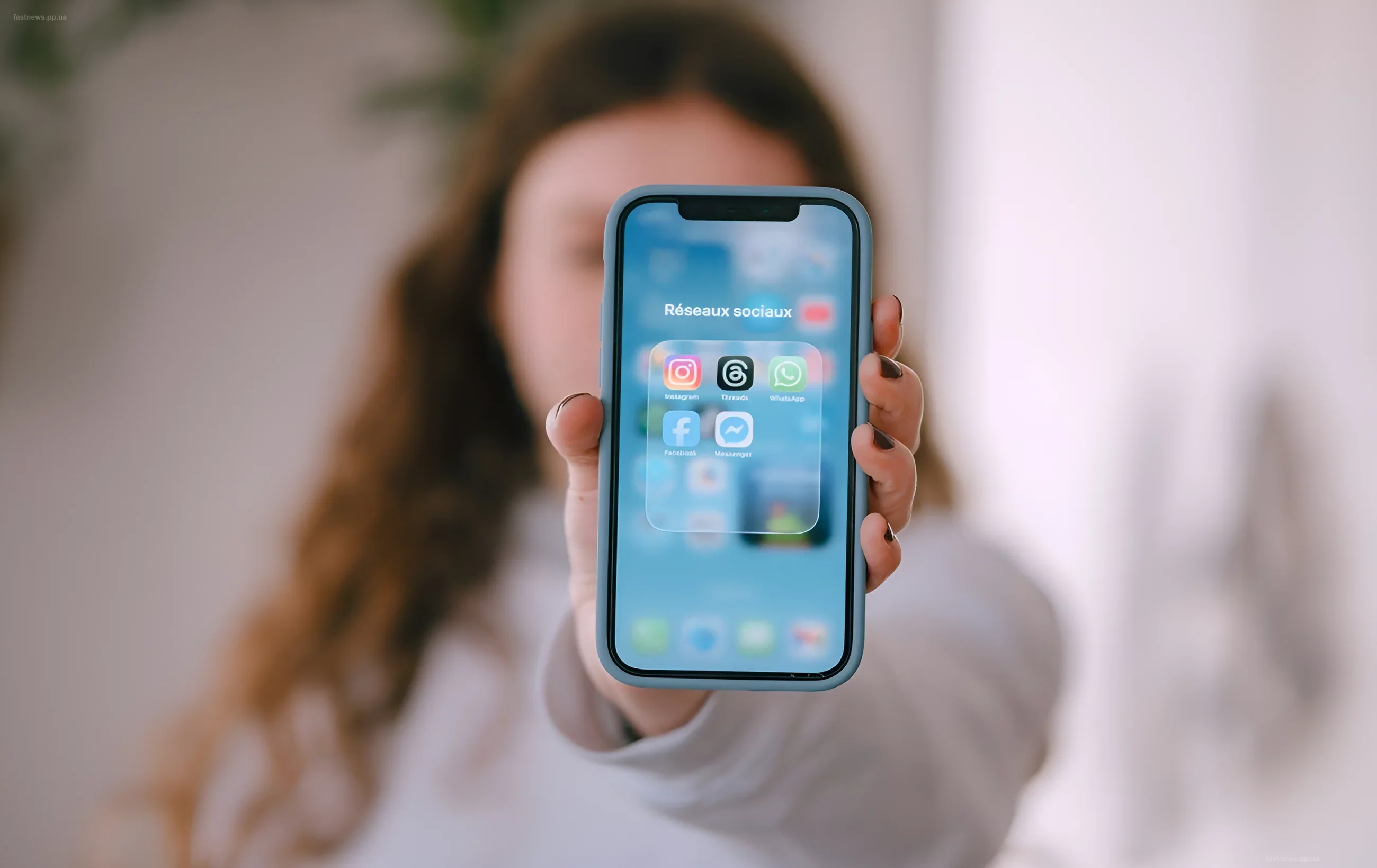 Женщина держит смартфон с открытым экраном, на котором видны иконки Instagram, Facebook и WhatsApp в папке "Социальные сети"