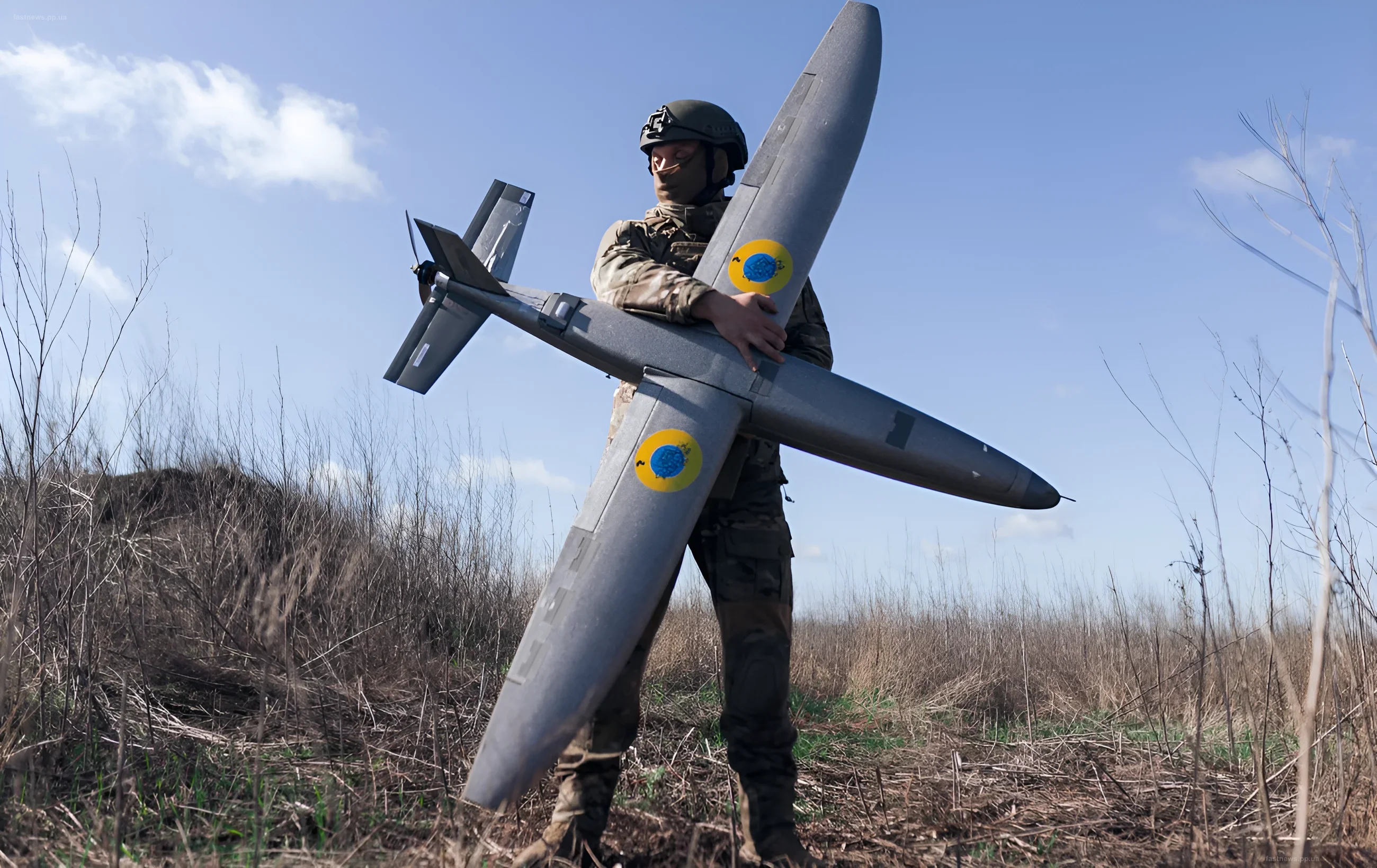 Украинский военный в камуфляже и шлеме держит в руках большой разведывательно-ударный дрон с опознавательными знаками Украины на фоне голубого неба и сухой травы.