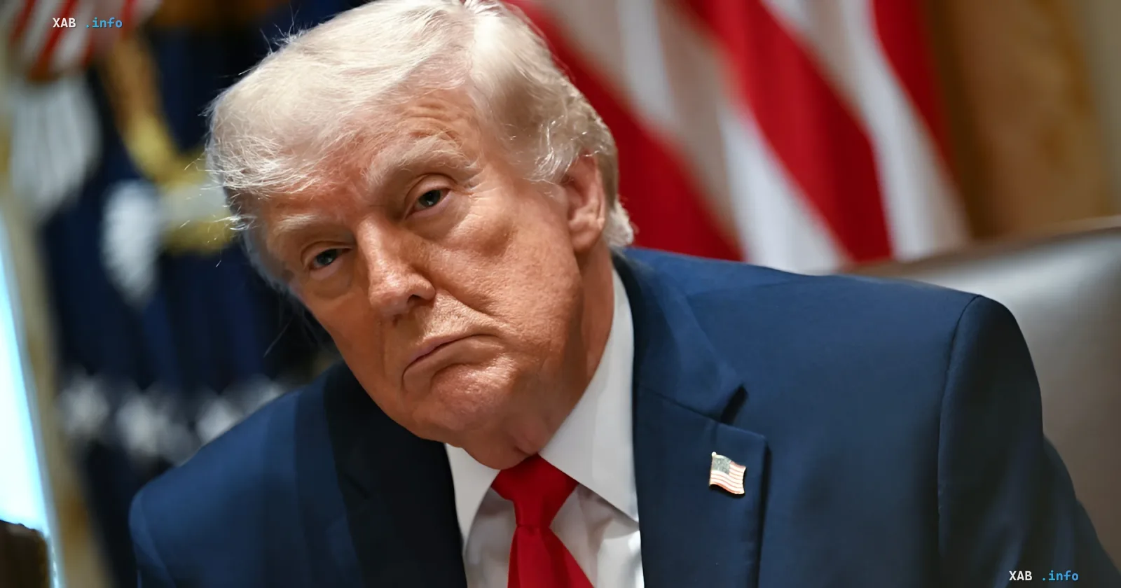 Дональд Трамп в синем костюме с красным галстуком и флагом США на лацкане, задумчиво смотрит в сторону, на фоне американский флаг — иллюстрация к новости о требовании пересмотра ядерной сделки с Ираном