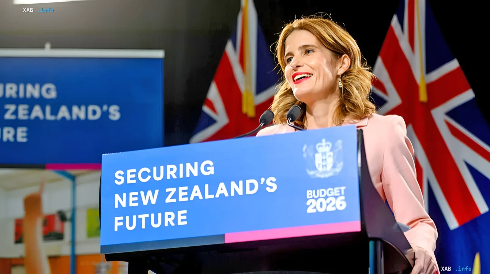 Neuseeländische Finanzministerin an einem Podium mit dem Slogan 'SECURING NEW ZEALAND'S FUTURE' und der Aufschrift 'BUDGET 2026', was die Antwort des Landes auf die US-Forderung nach erhöhten Militärausgaben symbolisiert