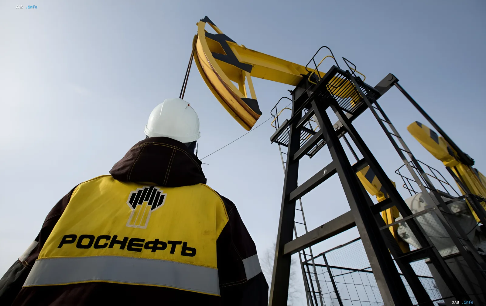 Работник «Роснефти» в защитной форме и каске стоит перед нефтяной вышкой, символизируя влияние 21-го пакета санкций ЕС на российскую нефтегазовую отрасль.