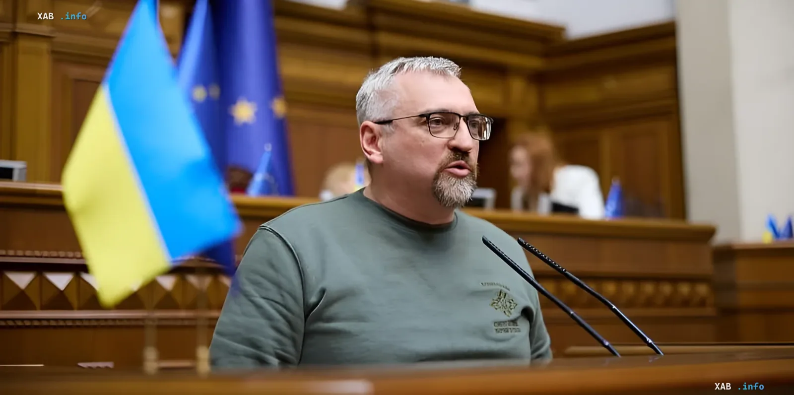 Man in green shirt with emblem stands at podium with Ukrainian and EU flags in background — possibly a legislator or official discussing the draft evaders registry