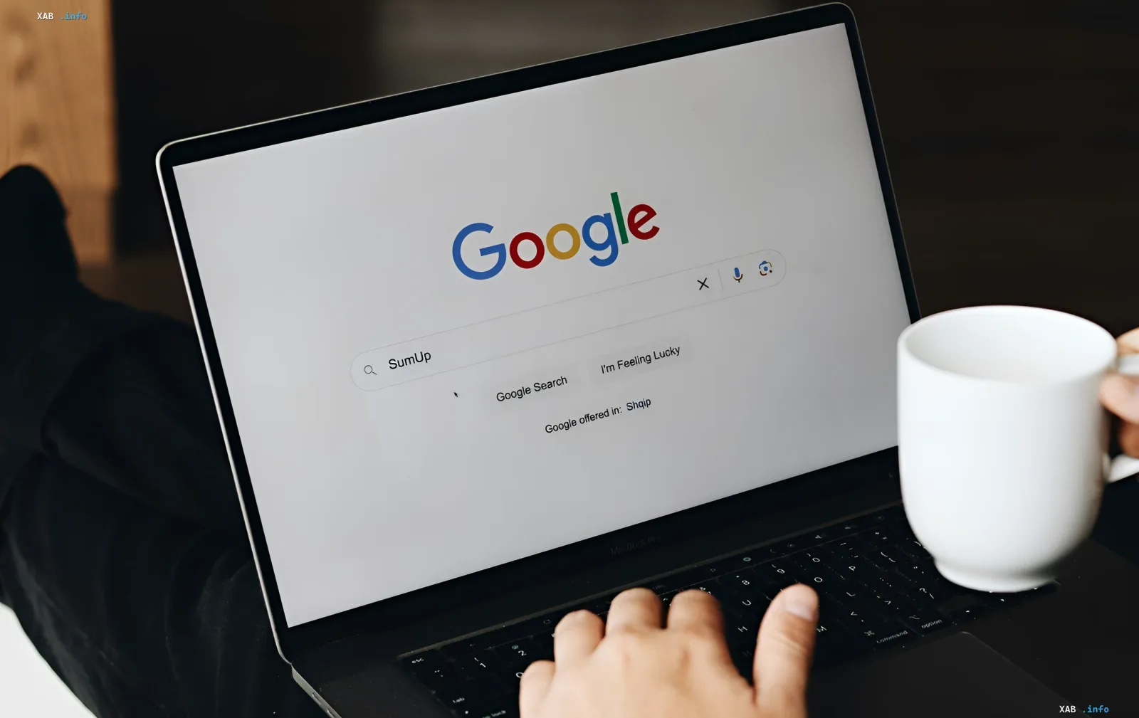 Eine Person in dunkler Kleidung sitzt mit einem Laptop, auf dessen Bildschirm die Google-Startseite mit der Suchanfrage 'SumUp' angezeigt wird, daneben steht eine weiße Kaffeetasse — Illustration zur Nachricht, dass Google Websites nun erlaubt, ihren Inhalt aus der KI-Suche zu blockieren