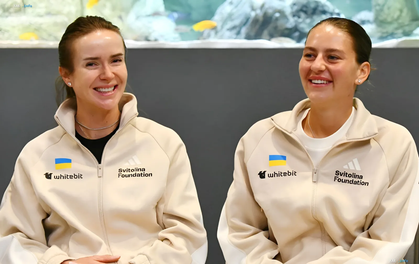 Элина Свитолина и Марта Костюк в одинаковых куртках с логотипами Svitolina Foundation и Whitebit, улыбаются на фоне аквариума — символ их успеха и выхода в топ-8 WTA