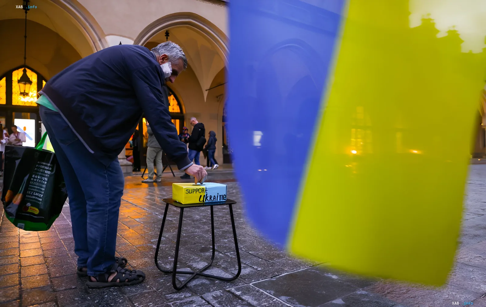 Чоловік кидає монету в скриньку з написом 'Support Ukraine' на тлі українського прапора, що символізує підтримку в умовах посилення правил в'їзду для призовників