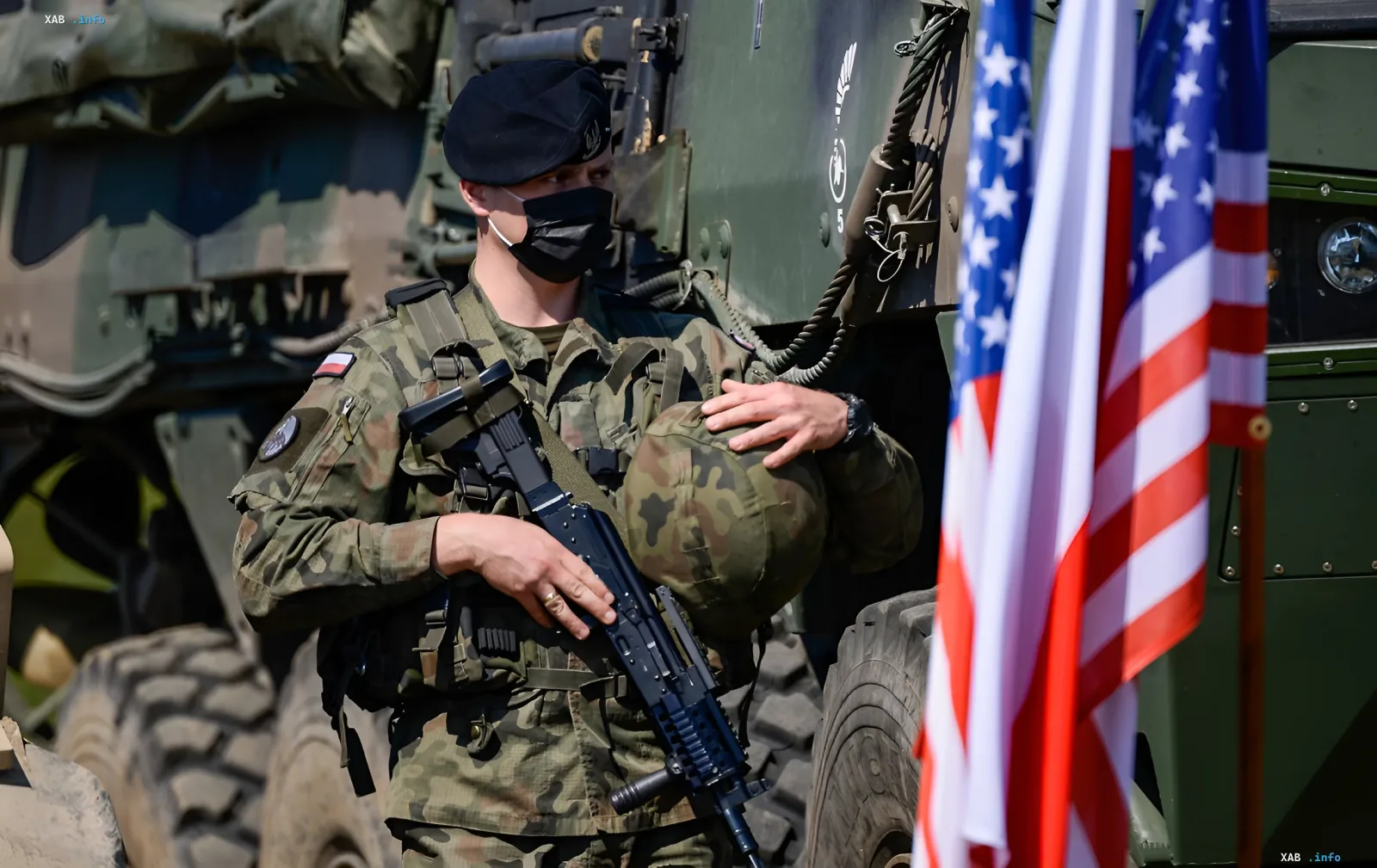 Американский солдат в маске с оружием стоит у военной техники на фоне флагов США и Польши, иллюстрируя эскалацию конфликта после ударов по Ирану