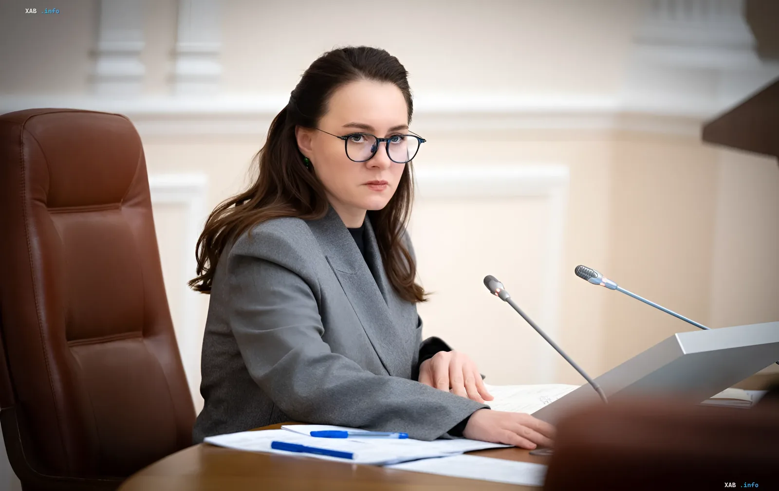 Женщина-чиновник за столом с документами и микрофонами, символизирующая принятие новых правил поддержки переселенцев и семей с детьми в 2026 году