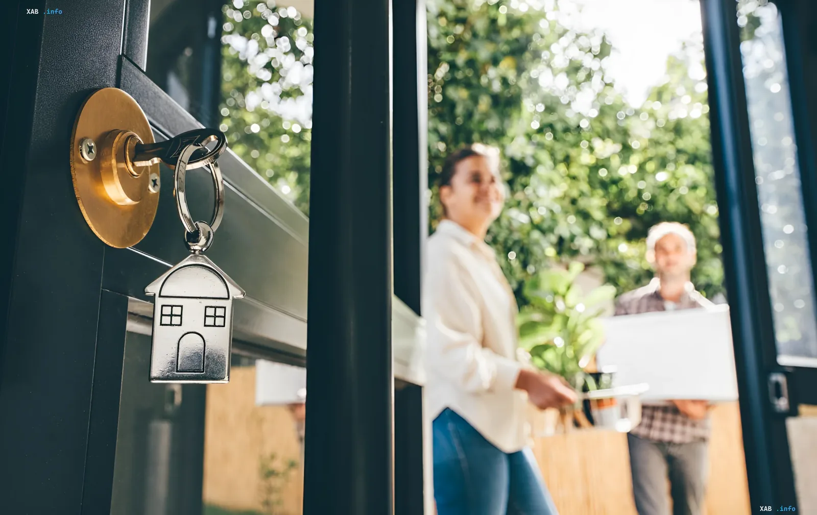 Key with a house-shaped keychain in the door lock, in the background a happy couple with boxes moves into a new apartment, illustrating the legalization of housing rental in Ukraine