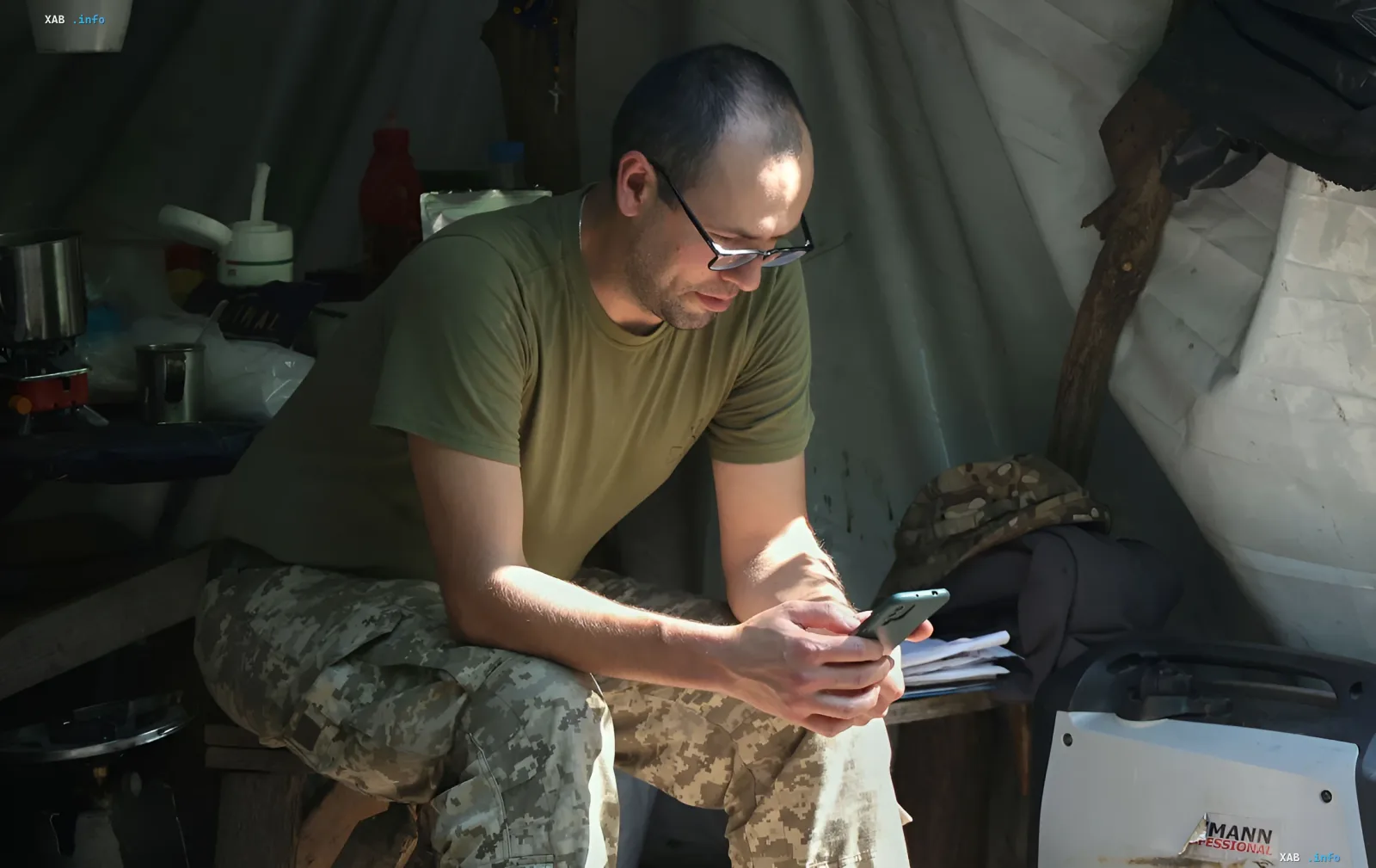 A Ukrainian soldier in camouflage uniform sits in a field shelter and looks at his smartphone, possibly checking information about salary or a new contract.