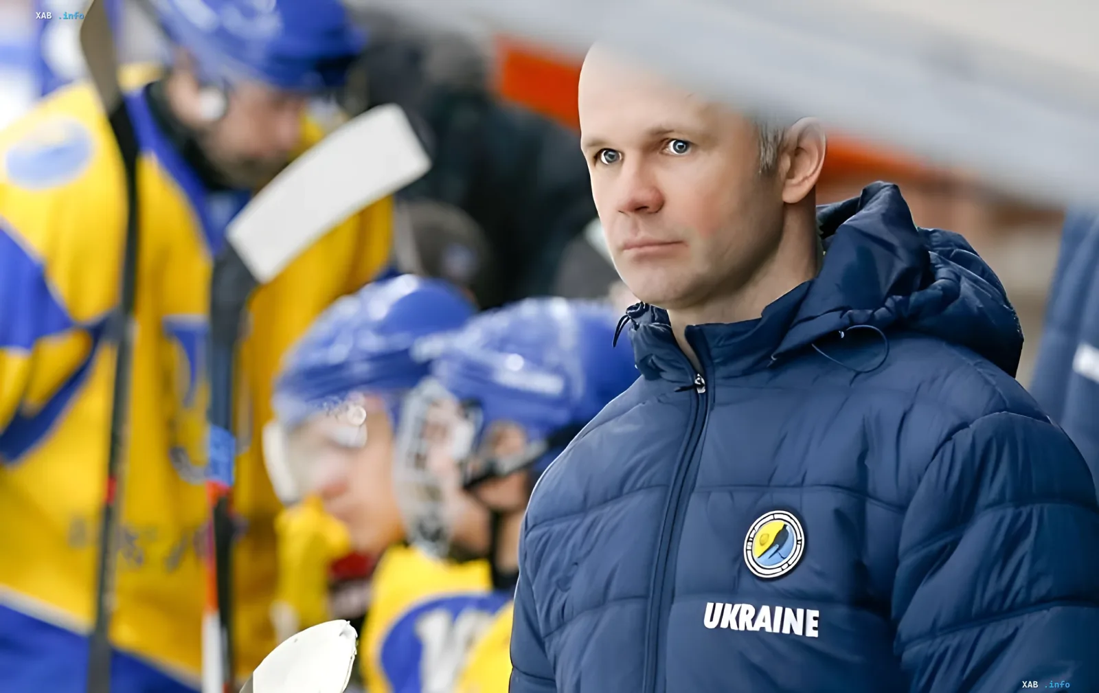Головний тренер збірної України з хокею Андрій Срюбко в синій куртці з логотипом України на фоні гравців у жовто-синіх формах
