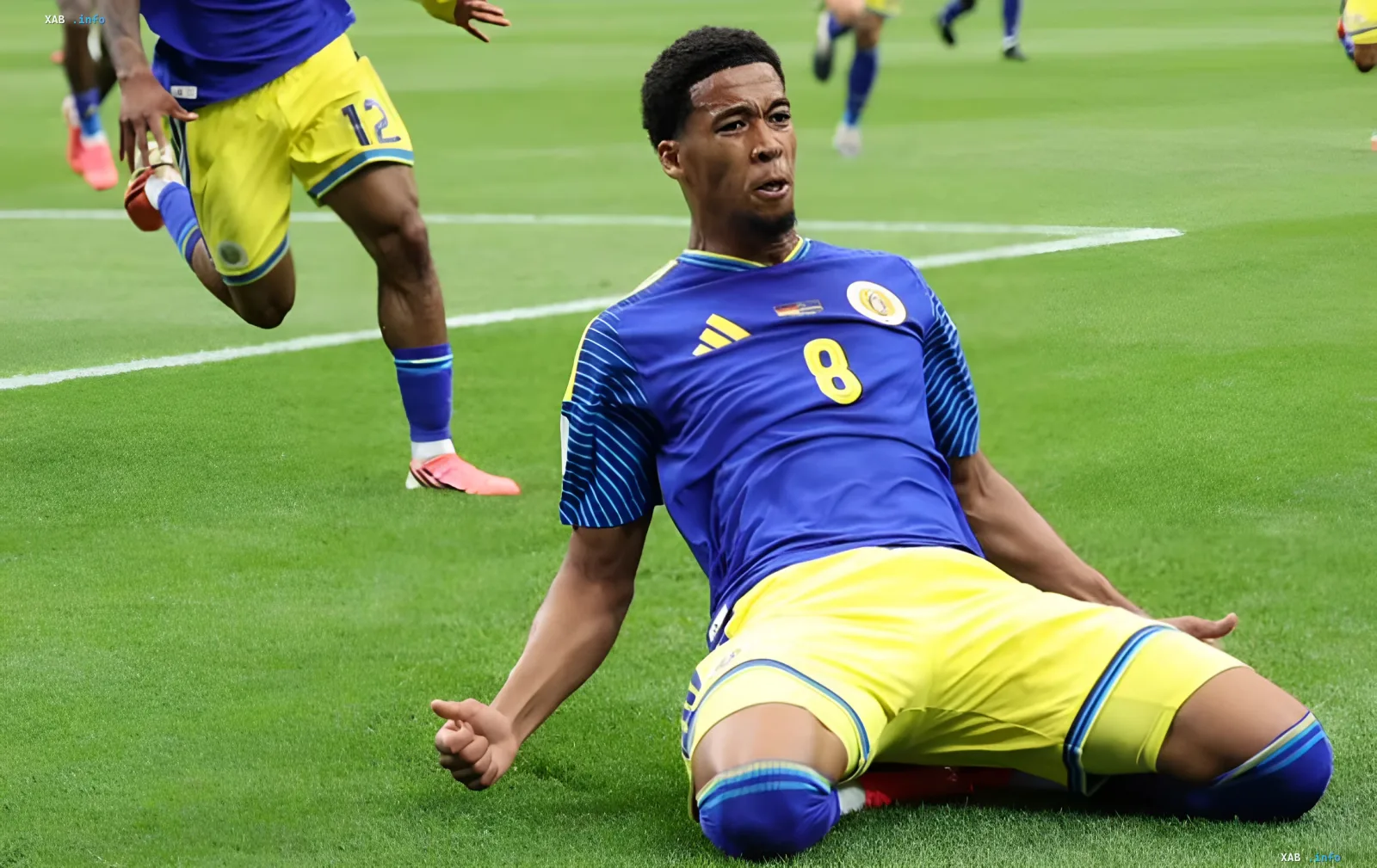 Curaçao football player in a blue jersey with yellow shorts and number 8 sits on the pitch, expressing disappointment after a crushing defeat by Germany at the 2026 World Cup