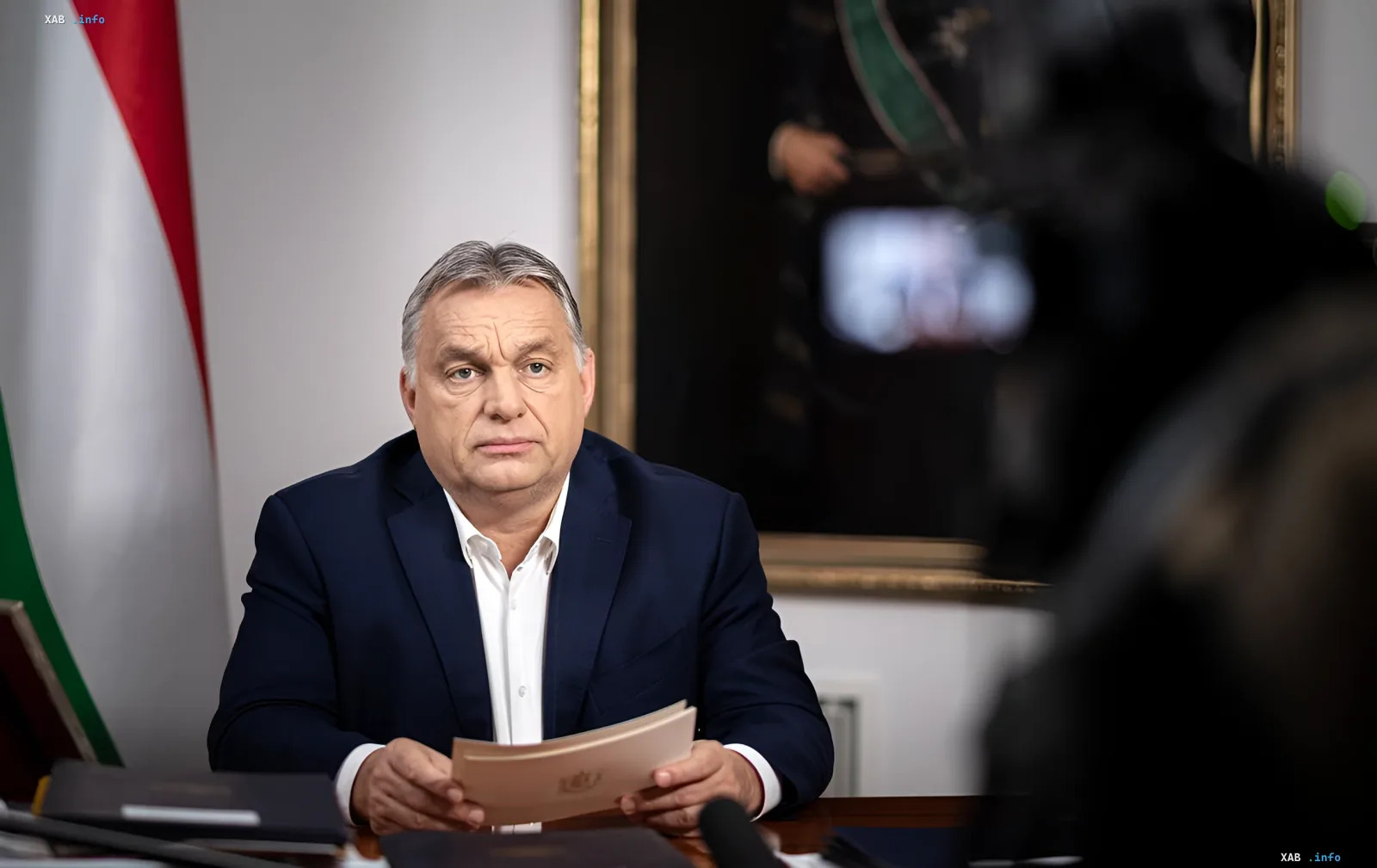 Виктор Орбан, премьер-министр Венгрии, сидит за столом с документами в руках, на фоне венгерский флаг. Символизирует конец его эпохи власти после конституционного запрета.