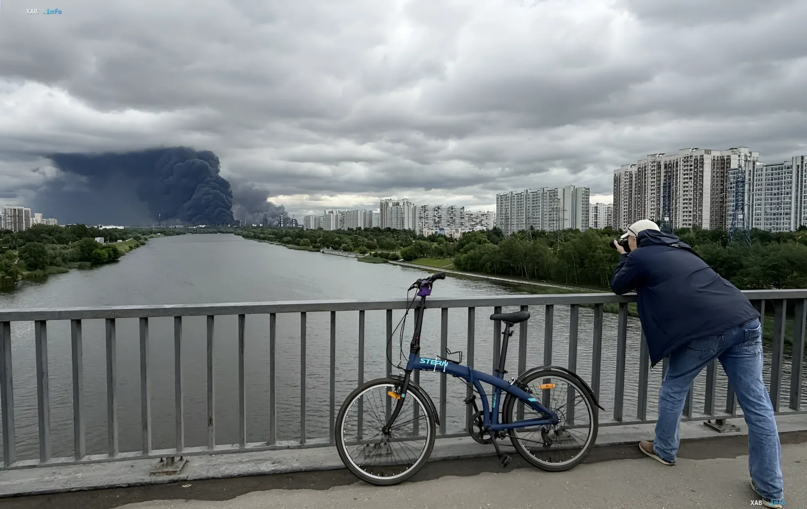 Photographer captures thick black smoke over Moscow from a bridge, with a bicycle nearby and modern residential buildings in the background