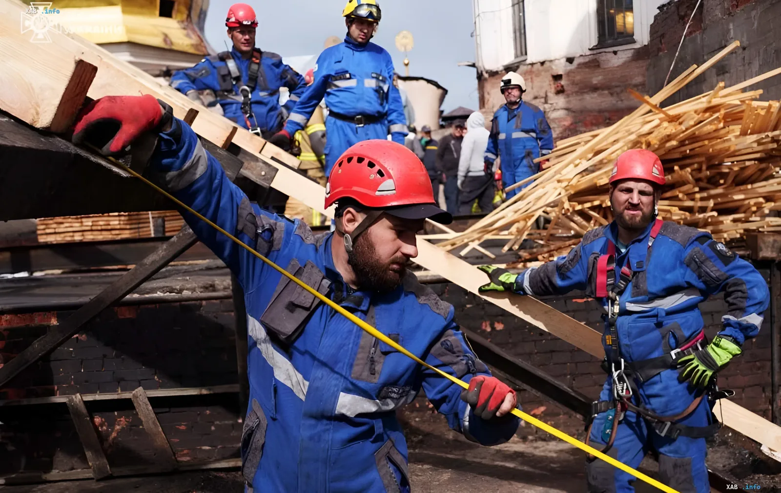 Rescue workers in blue uniforms and red helmets restore the wooden roof structure of the Assumption Cathedral after shelling, working with beams and construction materials.